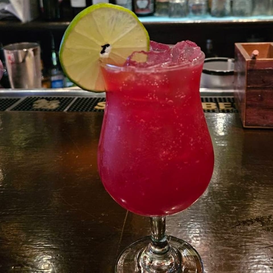 A vibrant red cocktail with ice and a lime wedge sits on a dark bar counter.