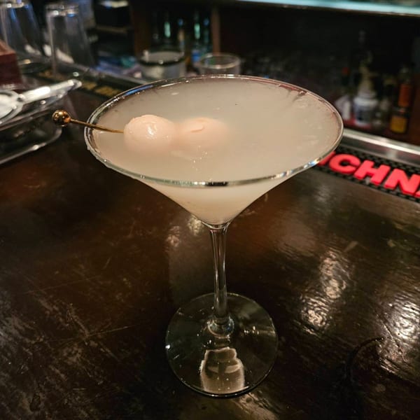 A lychee martini with two lychees on a skewer, sitting on a dark wooden bar.