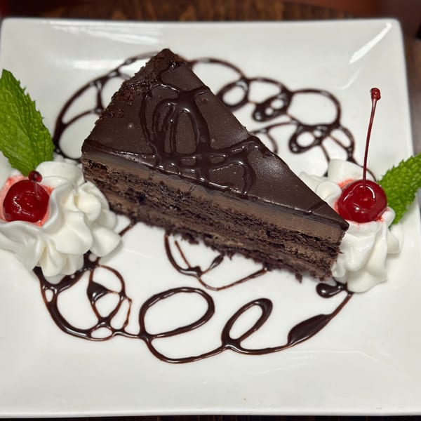 A slice of rich chocolate cake with chocolate drizzle, whipped cream, cherries, and mint on a white plate.