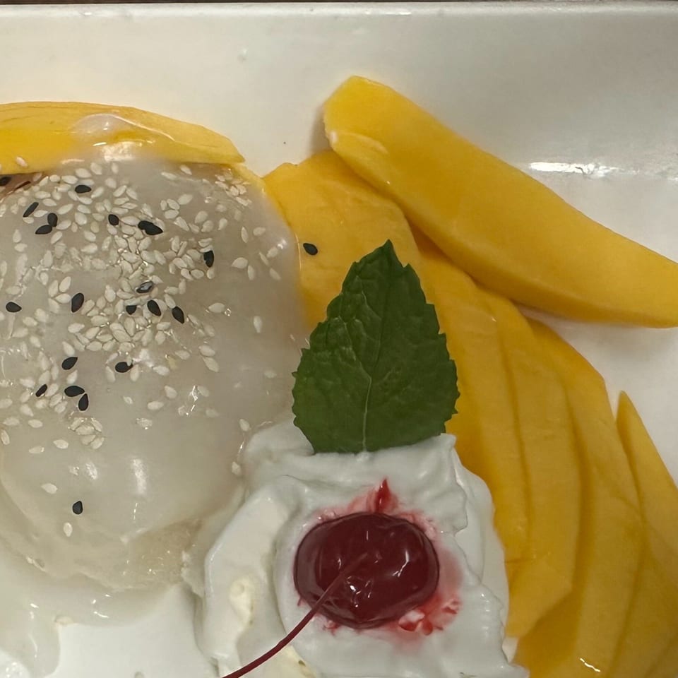 Erawan's mango sticky rice dessert with whipped cream, a cherry, and mint on a white plate.
