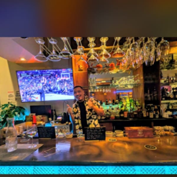 A smiling bartender in a Hawaiian shirt gives a thumbs-up behind a well-stocked bar at Erawan.