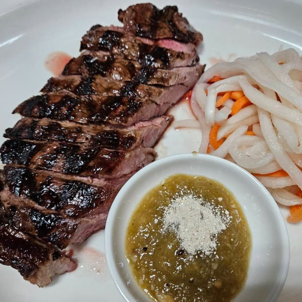 Sliced grilled steak with a side of pickled vegetables and a green dipping sauce on a white plate.