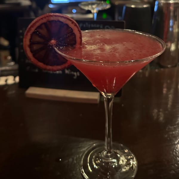 A vibrant red cocktail in a martini glass, garnished with a blood orange slice, on a dark bar.