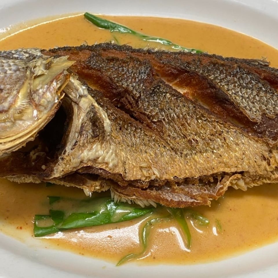 Crispy whole fried fish in a rich, creamy orange sauce with green vegetables on a white plate.