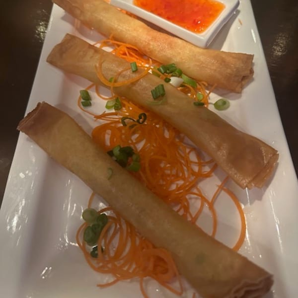 Three crispy Thai spring rolls on a white plate, garnished with shredded carrots and green onions, with dipping sauce.