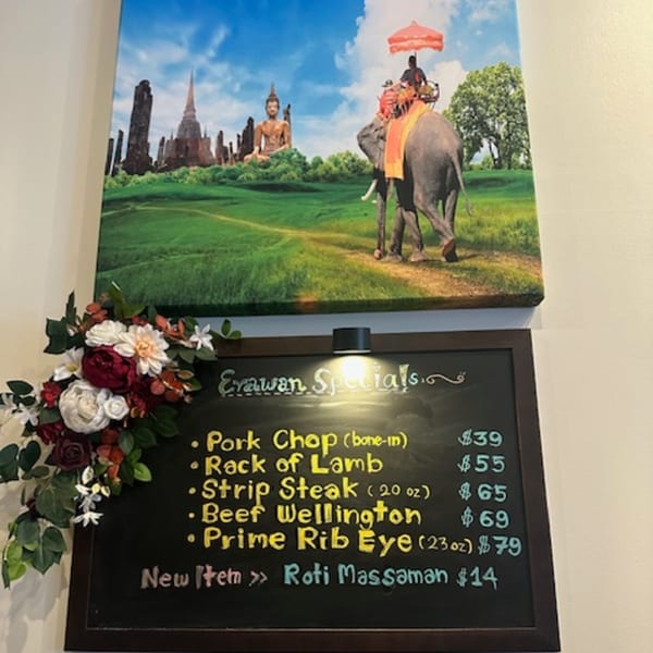 Erawan Specials chalkboard menu with prices, adorned with flowers, beneath a Thai landscape painting.